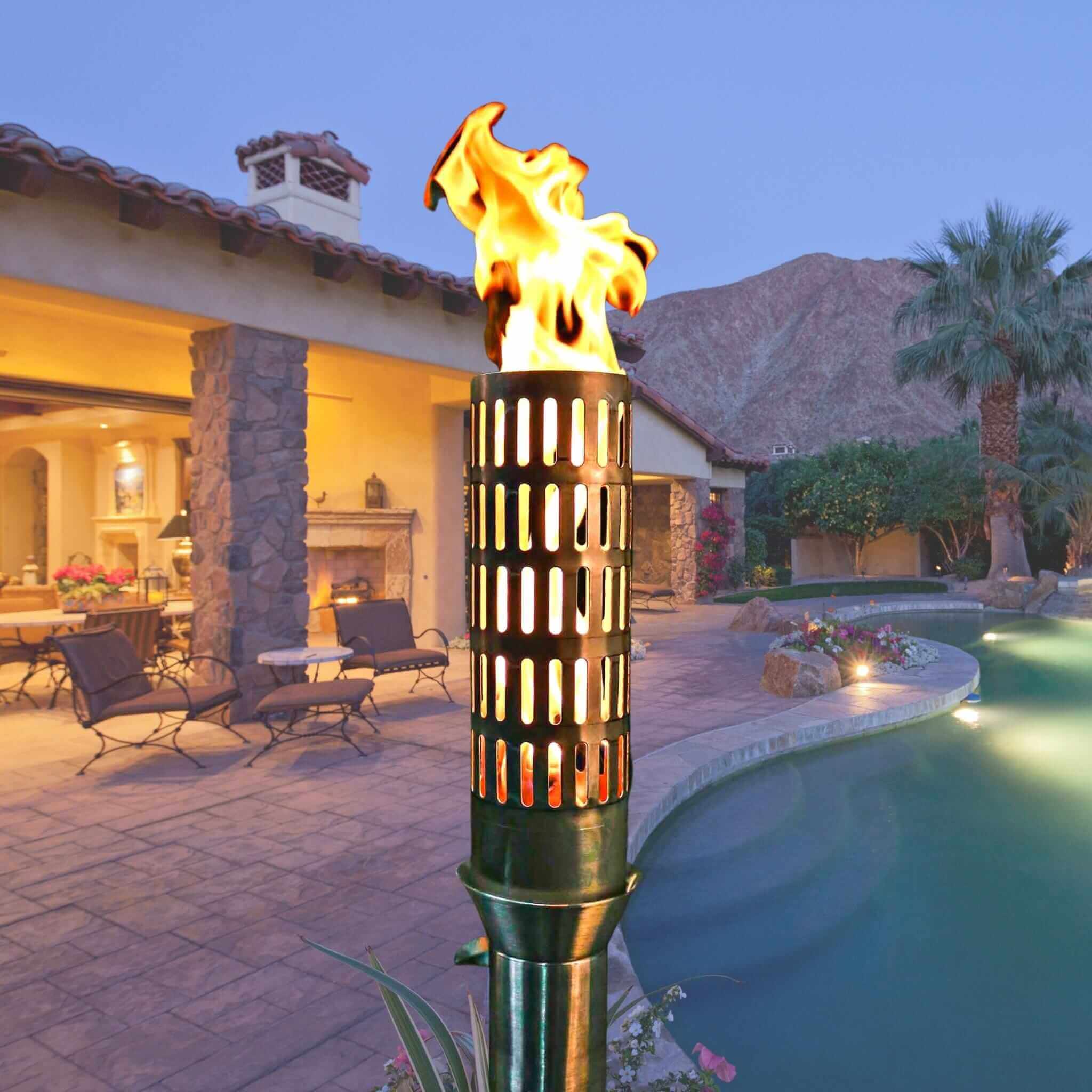 Flaming outdoor torch in a backyard setting with pool and house in the background. The Vent Fire Torch is a sculptural, stainless-steel gas torch designed to elevate upscale outdoor environments. At 14″ tall, its vented barrel emits a soft, inviting glow while providing ambient warmth. Ideal for pathways, poolside settings, patios, or landscape accents, this handcrafted torch blends durability with aesthetic finesse.