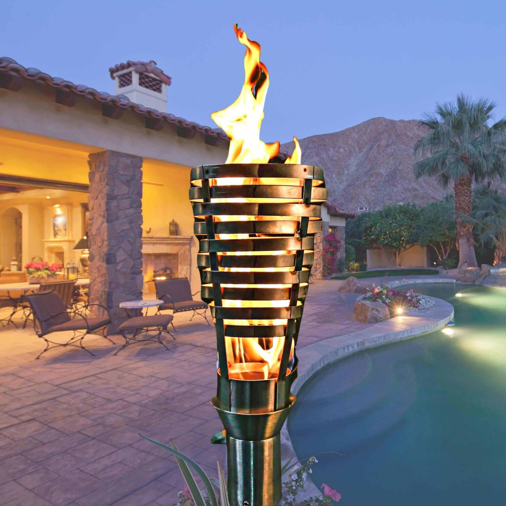 Decorative outdoor torch with flames in a backyard setting with pool and house. The Hercules Fire Torch by The Outdoor Plus is a striking outdoor statement piece, handcrafted from premium 304 stainless steel. Inspired by classical designs and engineered for modern elegance, this torch stands at approximately 14″ tall with an 8″ diameter head. It creates a warm, flickering glow, ideal for lining paths, enhancing pool decks, or accentuating upscale landscaping.