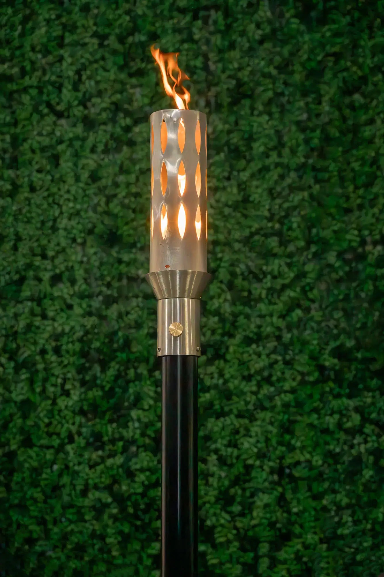 Torch with a burning flame against a green background.The Ellipse Fire Torch is a sleek, stainless-steel outdoor accent that marries contemporary elegance with responsive flame artistry. Standing about 14″ tall with a slender 3¾″ diameter, this torch elevates any outdoor space, patio edges, poolside perimeters, or garden pathways—with a warm, flickering signature flame.