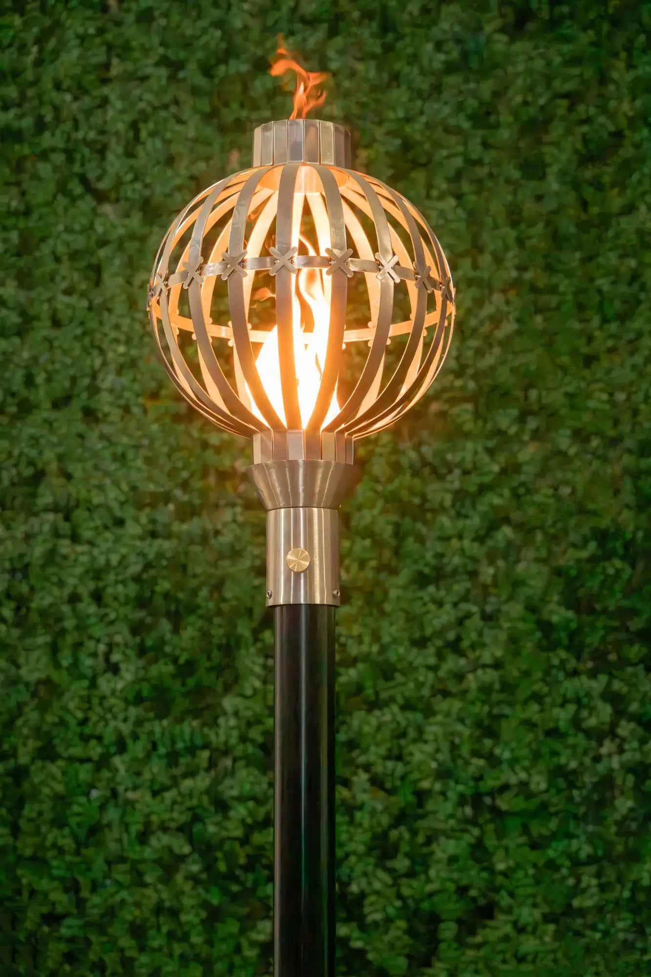 Decorative outdoor torch with a metal cage design against a green background. The Globe Fire Torch is a beautifully sculpted stainless-steel accent piece, handcrafted to elevate outdoor living spaces. Standing approximately 14" tall with an elegant spherical head, it disperses a warm, ambient glow across patios, pool perimeters, and garden paths. Its refined design brings a luxurious and modern aesthetic to any upscale environment.