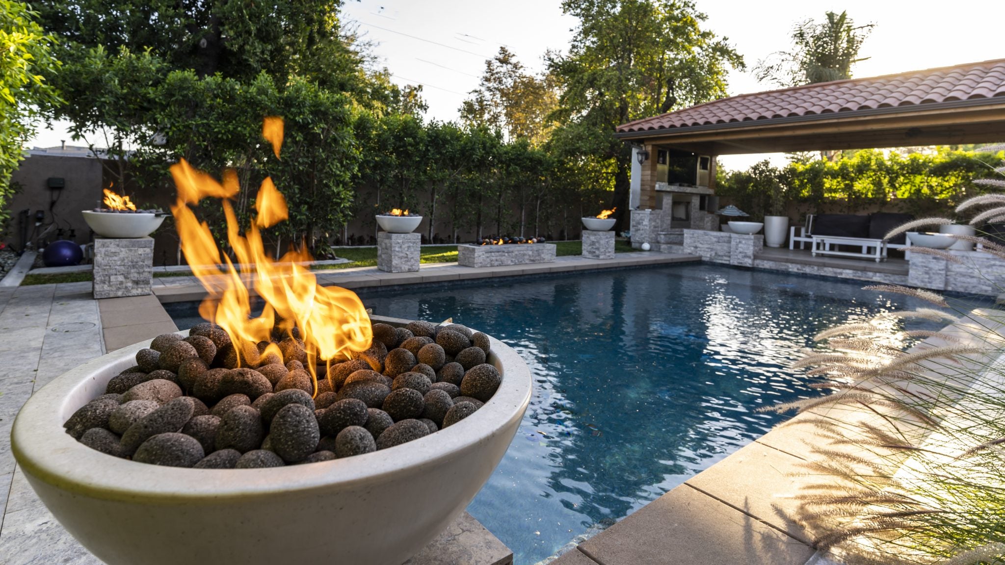 Fire pit with flames by a pool in a backyard setting. The Luna Concrete Fire Bowl is a beautifully modern, half-moon inspired fire feature designed to elevate outdoor spaces. Handcrafted in the USA from lightweight yet durable GFRC (Glass Fiber Reinforced Concrete), it delivers sculptural impact and cozy ambiance. Perfect for pool decks, patios, or urban rooftops.