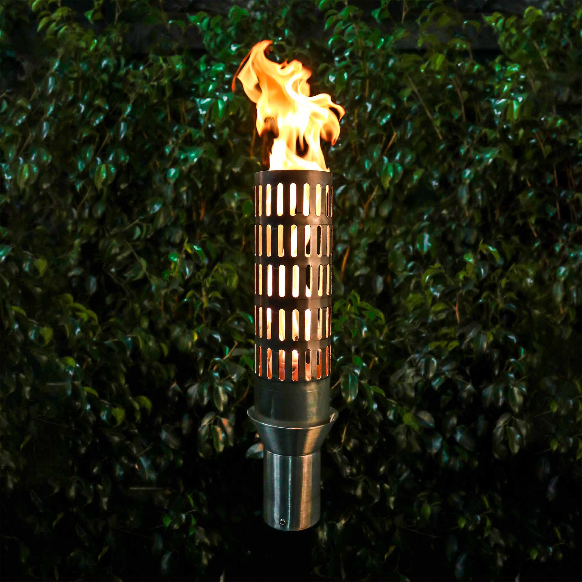 Tiki torch with flame against a green hedge background. The Vent Fire Torch is a sculptural, stainless-steel gas torch designed to elevate upscale outdoor environments. At 14″ tall, its vented barrel emits a soft, inviting glow while providing ambient warmth. Ideal for pathways, poolside settings, patios, or landscape accents, this handcrafted torch blends durability with aesthetic finesse.