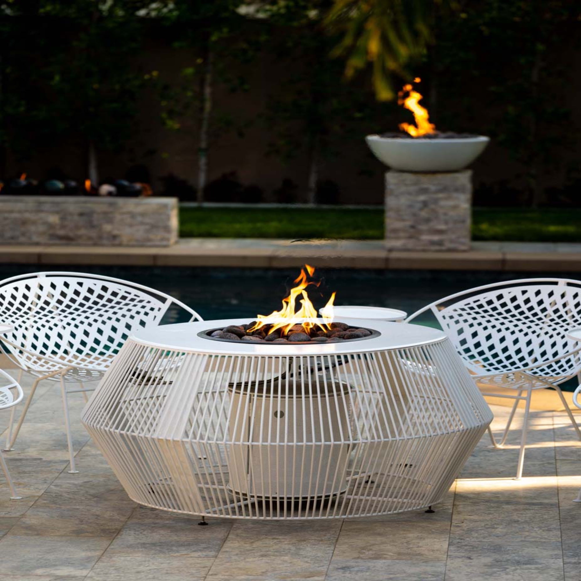 Outdoor patio setting with a fire pit table and chairs by a pool. The Cesto Fire Pit – Black &amp; White Collection combines modern form with refined outdoor living. With its minimalistic metal powder-coated finish in contrasting monochromatic tones, it anchors any patio, poolside, or lounge area while serving as both stylish furniture and functional fire feature.