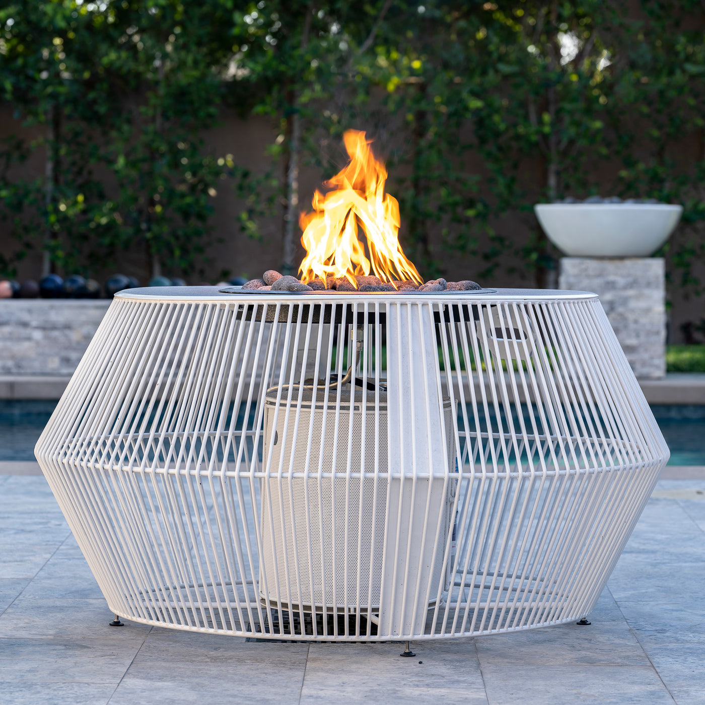 Outdoor fire pit table with flames in a backyard setting. The Cesto Fire Pit – Black &amp; White Collection combines modern form with refined outdoor living. With its minimalistic metal powder-coated finish in contrasting monochromatic tones, it anchors any patio, poolside, or lounge area while serving as both stylish furniture and functional fire feature.