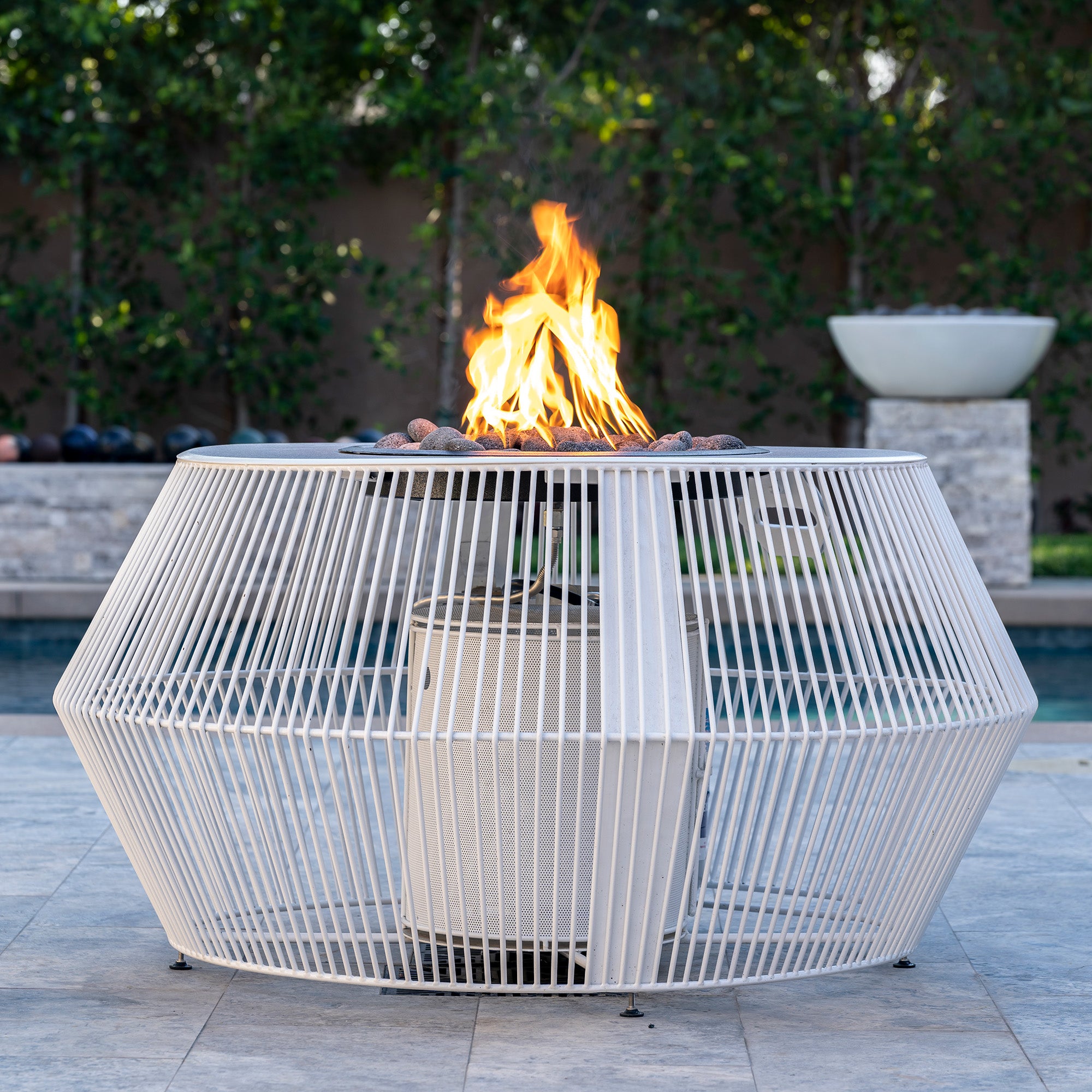 Outdoor fire pit table with flames in a backyard setting. The Cesto Fire Pit – Black &amp; White Collection combines modern form with refined outdoor living. With its minimalistic metal powder-coated finish in contrasting monochromatic tones, it anchors any patio, poolside, or lounge area while serving as both stylish furniture and functional fire feature.