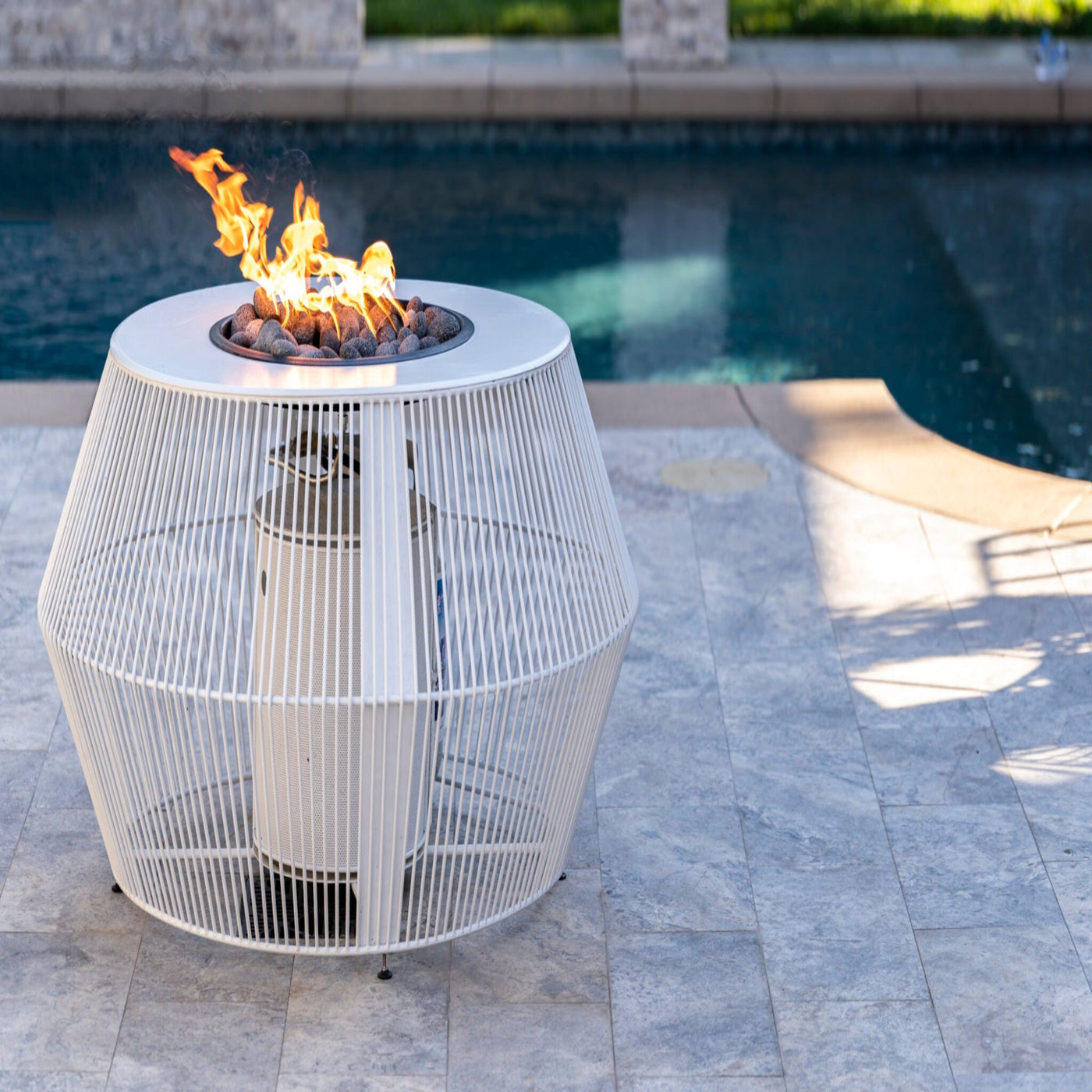 White wire fire pit table with flames by a pool. The Cesto Fire Pit – Black &amp; White Collection combines modern form with refined outdoor living. With its minimalistic metal powder-coated finish in contrasting monochromatic tones, it anchors any patio, poolside, or lounge area while serving as both stylish furniture and functional fire feature.
