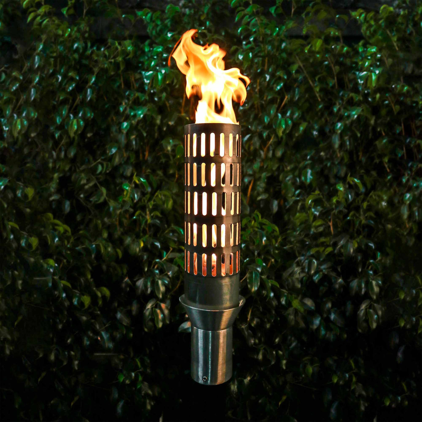 Tiki torch with flame against a green hedge background. The Vent Fire Torch is a sculptural, stainless-steel gas torch designed to elevate upscale outdoor environments. At 14″ tall, its vented barrel emits a soft, inviting glow while providing ambient warmth. Ideal for pathways, poolside settings, patios, or landscape accents, this handcrafted torch blends durability with aesthetic finesse.
